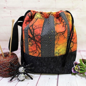 May include: A drawstring bag with a black handle and a striking orange and black tree branch pattern. The bag is paired with a ball of yarn, knitting needles, and a decorative spider. The bag's base is a dark gray fabric.