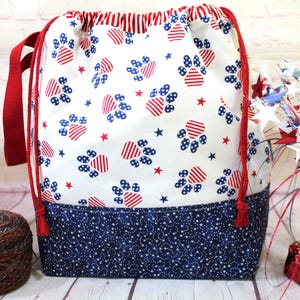May include: A drawstring project bag featuring a white fabric upper section with a red, white, and blue paw print and star pattern. The lower section is dark blue with a white star print. It includes red drawstring cords and red straps.