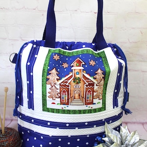 May include: A blue fabric tote bag featuring a central graphic of a gingerbread church surrounded by trees and stars. The bag has white polka dots on a blue background, a drawstring closure with blue cords, and two sturdy blue fabric handles.