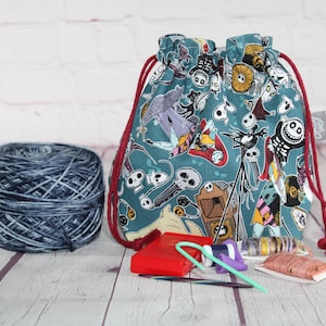 May include: A teal drawstring project bag with a Nightmare Before Christmas pattern, red drawstring, and a ball of blue and gray yarn. Knitting needles and other accessories are also visible.