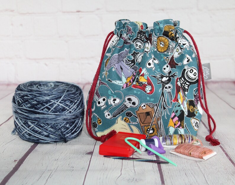 May include: A teal drawstring project bag with a Nightmare Before Christmas pattern, red drawstring, and a ball of blue and gray yarn. Knitting needles and other accessories are also visible.