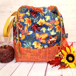 May include: A drawstring project bag featuring a blue fabric upper with a colorful autumn leaf and berry print, and a textured orange fabric base. The bag includes a yellow strap, brown rope drawstrings, and a small side tag.