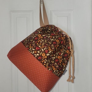 May include: A drawstring bag featuring a dark brown upper section with a pattern of autumn leaves, acorns, and mushrooms, and a burnt orange lower section with a white cross-stitch pattern. The bag has a beige strap and beige rope drawstrings.