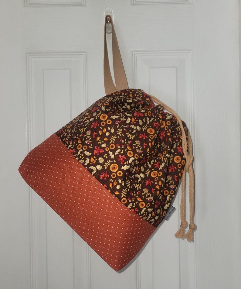 May include: A drawstring bag featuring a dark brown upper section with a pattern of autumn leaves, acorns, and mushrooms, and a burnt orange lower section with a white cross-stitch pattern. The bag has a beige strap and beige rope drawstrings.