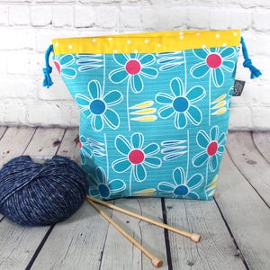 May include: A turquoise drawstring yarn bag featuring a floral pattern of blue and pink flowers. The bag has a yellow polka dot lining and blue drawstrings. A ball of blue yarn and two wooden knitting needles are in front.