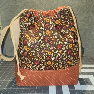 May include: A drawstring bag featuring a dark brown upper section with a repeating pattern of autumn leaves, mushrooms, and acorns. The lower section is a solid rust-orange fabric with a white polka dot pattern. It has beige rope straps.