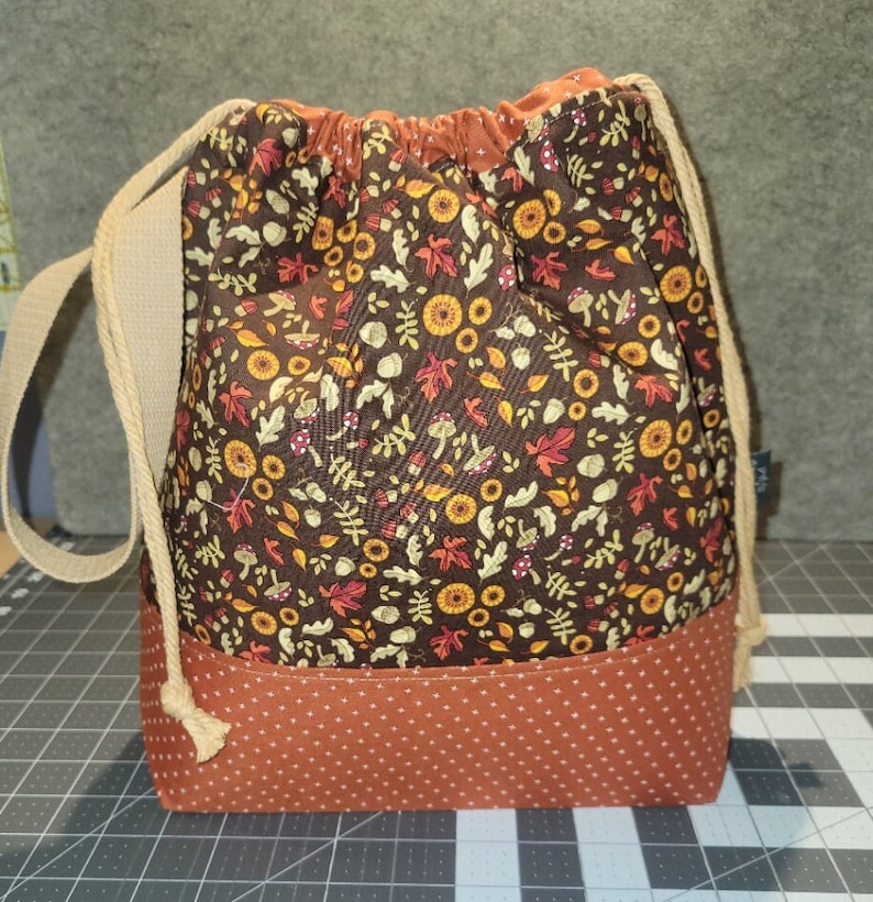 May include: A drawstring bag featuring a dark brown upper section with a repeating pattern of autumn leaves, mushrooms, and acorns. The lower section is a solid rust-orange fabric with a white polka dot pattern. It has beige rope straps.
