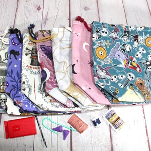 May include: A collection of drawstring project bags in various patterns, including skulls, cats, moons, and a teal Nightmare Before Christmas print. Accessories like a red stitch counter, needles, and yarn are also visible.