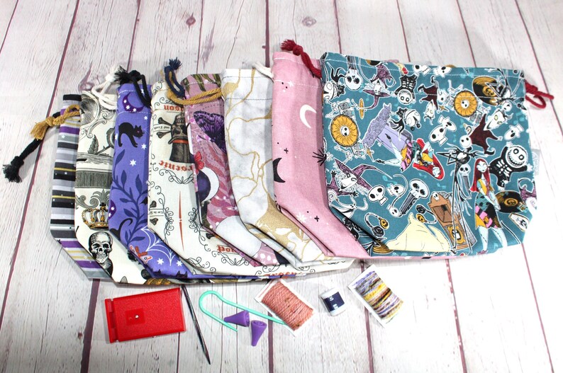 May include: A collection of drawstring project bags in various patterns, including skulls, cats, moons, and a teal Nightmare Before Christmas print. Accessories like a red stitch counter, needles, and yarn are also visible.