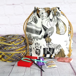 May include: A drawstring project bag featuring a white fabric with black and gold illustrations of skeletons, spiders, and clocks. Beside the bag sits a ball of variegated yellow, gray, and brown yarn, a red plastic case, and various sewing notions.