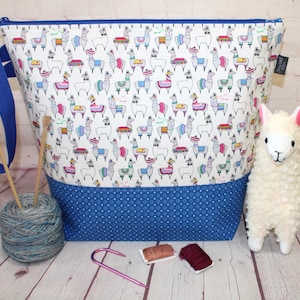 May include: A white fabric knitting project bag with a blue zipper and handles. The bag features a pattern of colorful llamas. A blue yarn ball, knitting needles, and a stuffed llama are next to the bag. Additional knitting supplies are also present.