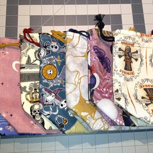 May include: A collection of drawstring bags with various Halloween-themed patterns. Designs include bats, skulls, moons, and the word "Poison". The bags are made of fabric and have drawstrings in various colors.