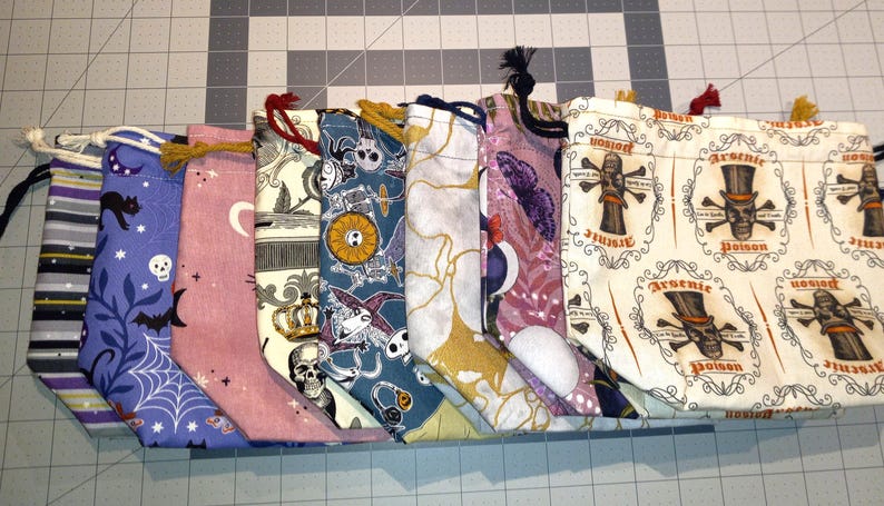 May include: A collection of drawstring bags with various Halloween-themed patterns. Designs include bats, skulls, moons, and the word "Poison". The bags are made of fabric and have drawstrings in various colors.