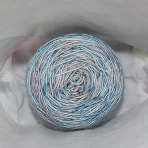 May include: A ball of yarn with a blend of blue, white, and pink fibers. The yarn is tightly wound and appears soft. The ball is nestled in a white fabric, suggesting a craft or knitting project.