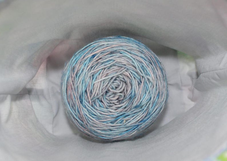 May include: A ball of yarn with a blend of blue, white, and pink fibers. The yarn is tightly wound and appears soft. The ball is nestled in a white fabric, suggesting a craft or knitting project.