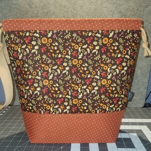 May include: A drawstring bucket bag featuring a dark brown main panel with a repeating pattern of autumn leaves, mushrooms, and acorns. The top and bottom sections are made of a rust-colored fabric with a small white cross-stitch pattern.