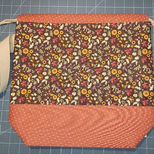 May include: A drawstring bag featuring a dark brown fabric panel with a repeating pattern of autumn leaves, mushrooms, and acorns, contrasted with a solid orange fabric base with a white cross-stitch pattern and a beige woven shoulder strap.