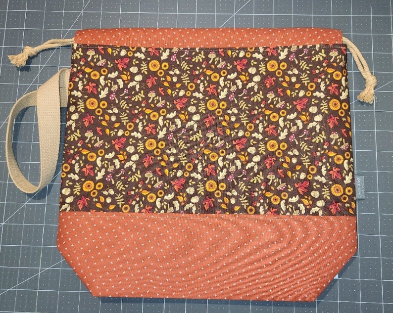 May include: A drawstring bag featuring a dark brown fabric panel with a repeating pattern of autumn leaves, mushrooms, and acorns, contrasted with a solid orange fabric base with a white cross-stitch pattern and a beige woven shoulder strap.