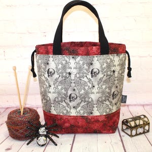 May include: A tote bag with a black handle and a white and gray skull and floral pattern. The top and bottom of the bag are red. A ball of yarn, knitting needles, and a decorative spider are next to the bag.