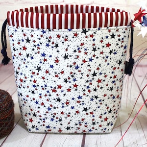 Puede incluir: Una bolsa de tela con cordón que presenta un exterior blanco con un patrón denso de pequeñas estrellas rojas y azules. El forro interior es de tela a rayas verticales rojas y blancas. La bolsa incluye cordones azul oscuro a los lados.