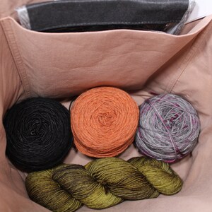 Puede incluir: Cuatro ovillos de lana en una bolsa de tela beige. La lana es en tonos de negro, naranja, verde y gris.