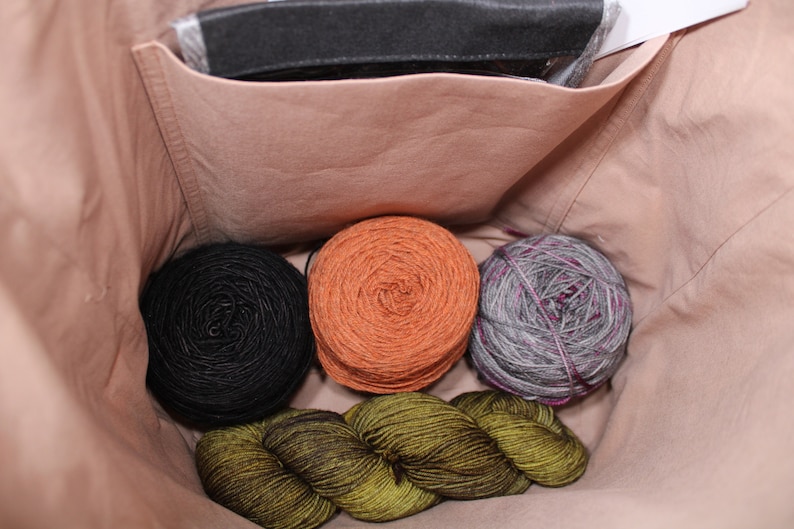 Puede incluir: Cuatro ovillos de lana en una bolsa de tela beige. La lana es en tonos de negro, naranja, verde y gris.