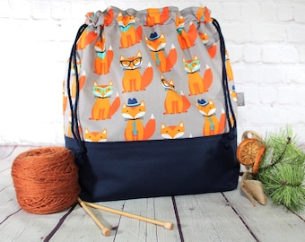 May include: A drawstring bag featuring a gray background with a repeating pattern of orange foxes wearing blue hats, ties, and glasses. The bottom section of the bag is solid navy blue, and it includes dark blue rope drawstrings at the top.