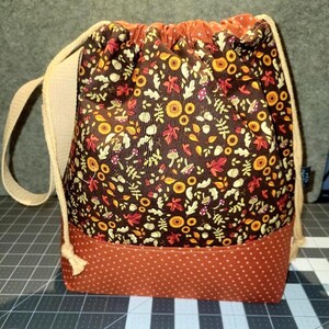May include: A drawstring bag featuring a dark brown fabric with a repeating pattern of yellow sunflowers, red maple leaves, and white mushrooms. The base is a solid burnt orange fabric with small white polka dots, and it has beige rope straps.