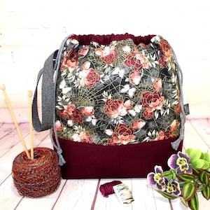 Puede incluir: Una bolsa de proyecto de tejido con cordón con un estampado floral de rosas rojas y telarañas doradas sobre un fondo negro. La bolsa tiene una base burdeos y un asa gris. Una madeja de hilo y agujas de tejer están al lado.
