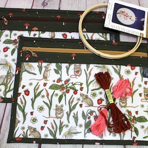 Könnte beinhalten: Zwei grüne und weiße Stoffbeutel mit einem Waldmaus- und Erdbeermuster, mit Reißverschluss. Ein hölzerner Stickrahmen, Stickgarn und ein Kreuzstichmuster sind ebenfalls vorhanden. Die Beutel sind für die Aufbewahrung von Bastelarbeiten konzipiert.