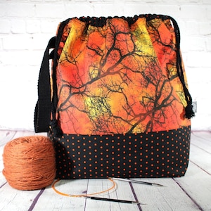 May include: A drawstring project bag featuring an orange and yellow sunset-colored fabric with a black tree branch silhouette pattern. The base is black with small orange polka dots. It includes black cord drawstrings and a black shoulder strap.