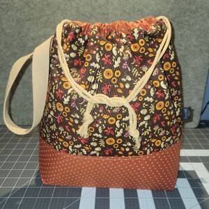 May include: A drawstring bag featuring a brown fabric with a pattern of autumn leaves, mushrooms, and flowers. The base is a solid burnt orange fabric with a white dot pattern. It includes a beige woven shoulder strap and beige rope ties.