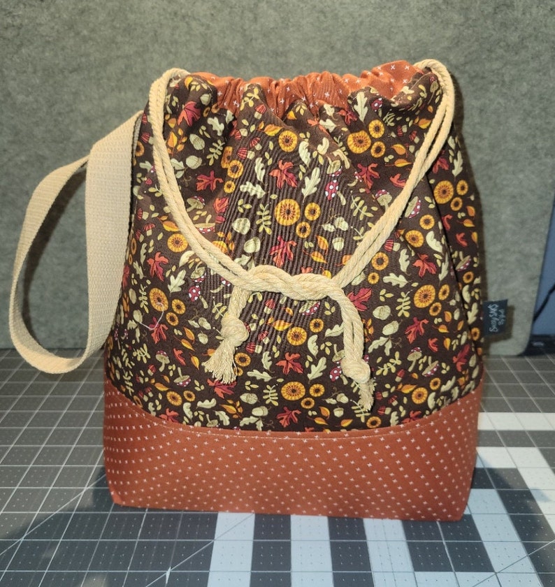 May include: A drawstring bag featuring a brown fabric with a pattern of autumn leaves, mushrooms, and flowers. The base is a solid burnt orange fabric with a white dot pattern. It includes a beige woven shoulder strap and beige rope ties.