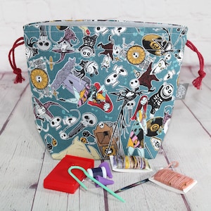 May include: A teal drawstring project bag featuring a repeating pattern of characters from The Nightmare Before Christmas. The bag has red drawstrings and a gray interior. Knitting accessories, including needles and yarn, are visible in front of the bag.