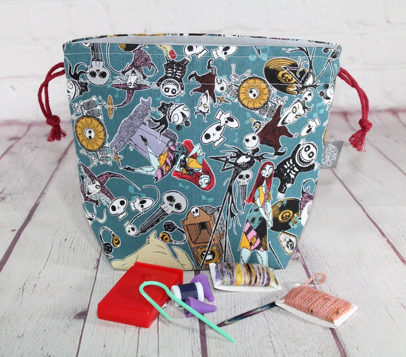 May include: A teal drawstring project bag featuring a repeating pattern of characters from The Nightmare Before Christmas. The bag has red drawstrings and a gray interior. Knitting accessories, including needles and yarn, are visible in front of the bag.