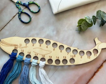 May include: Wooden whale-shaped thread organizer with 20 numbered holes for storing embroidery floss. The organizer is made of light brown wood and has a natural finish. Five skeins of blue and white embroidery floss are threaded through the holes.