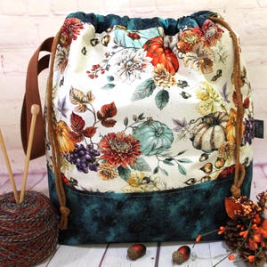 May include: A drawstring project bag featuring a cream-colored fabric with a print of pumpkins, flowers, and grapes, contrasted by a dark teal bottom panel. The bag includes brown cord drawstrings and a brown fabric shoulder strap.