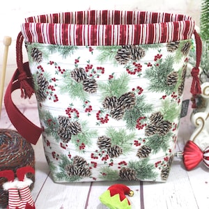 May include: A fabric drawstring project bag featuring a white background with a pattern of green pine branches, brown pinecones, and red berries. The bag has a dark red and white striped interior lining and matching dark red drawstring cords.