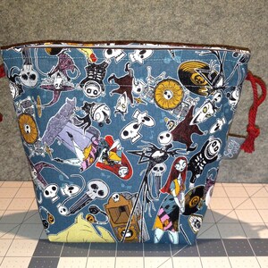 May include: A blue drawstring bag featuring a repeating pattern of characters and imagery from The Nightmare Before Christmas. The bag has red drawstrings and a brown trim. A small tag with text is attached to the side.