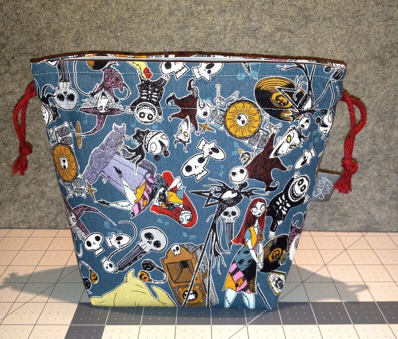 May include: A blue drawstring bag featuring a repeating pattern of characters and imagery from The Nightmare Before Christmas. The bag has red drawstrings and a brown trim. A small tag with text is attached to the side.