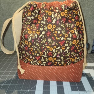 May include: A drawstring bag featuring a brown fabric upper section with a pattern of autumn leaves, mushrooms, and acorns, and a solid rust-colored fabric lower section with a white polka dot print. The bag includes beige rope straps.