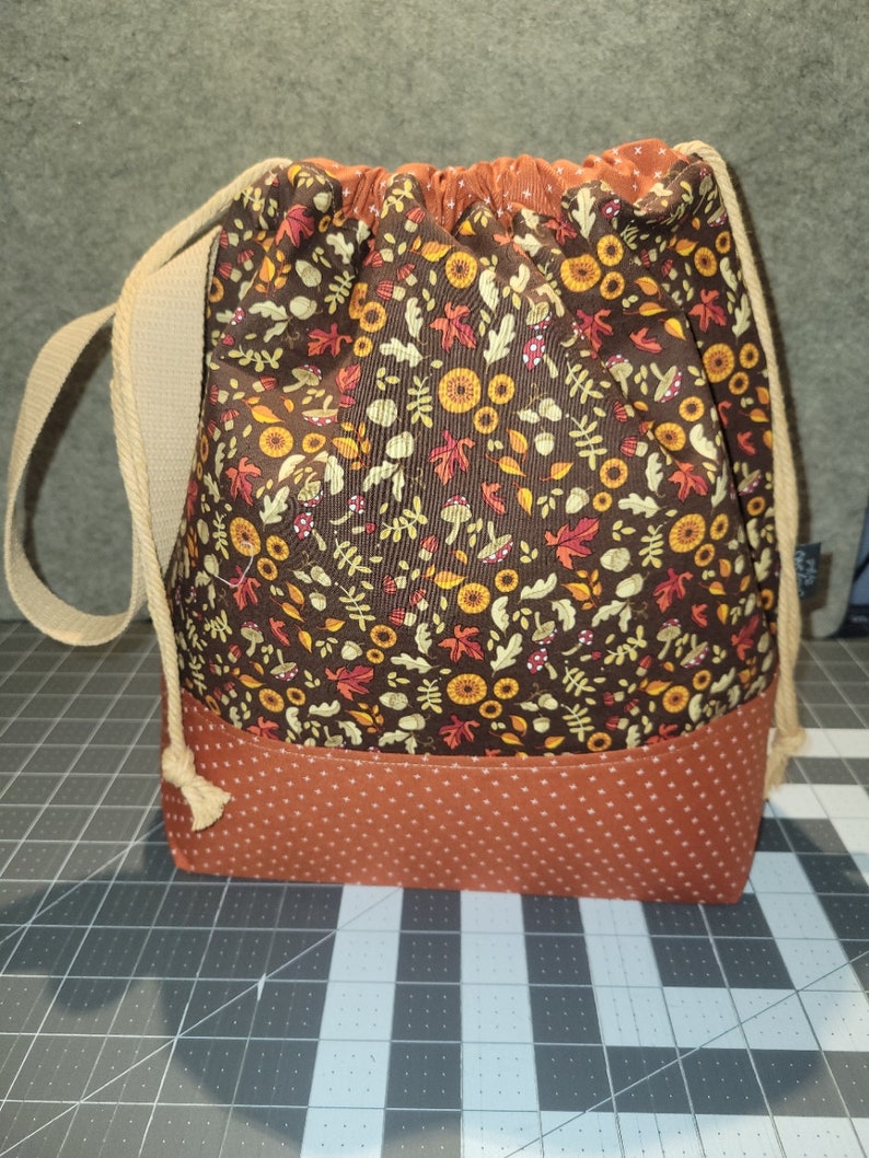 May include: A drawstring bag featuring a brown fabric upper section with a pattern of autumn leaves, mushrooms, and acorns, and a solid rust-colored fabric lower section with a white polka dot print. The bag includes beige rope straps.