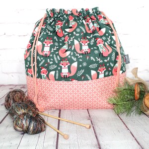 May include: A drawstring project bag featuring a dark green upper section with a repeating pattern of foxes wearing scarves and hats, and a light pink lower section with a geometric print. The bag has pink cord drawstrings and a small side tag.