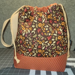 May include: A drawstring bag featuring a dark brown fabric upper section with a repeating pattern of autumn leaves, acorns, and mushrooms, and a solid burnt orange fabric lower section with a small white cross-stitch pattern and beige rope straps.