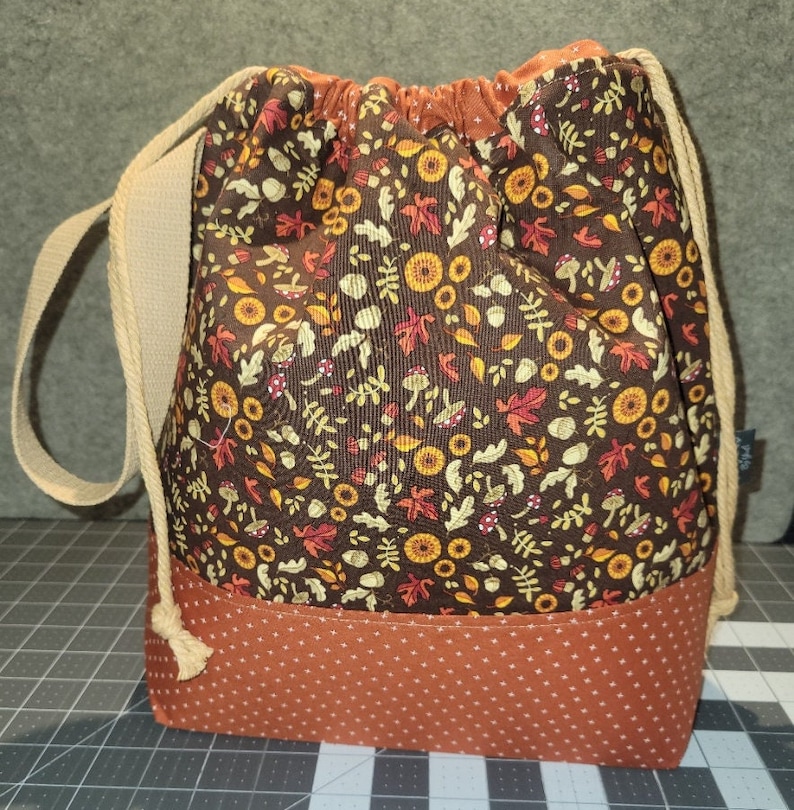 May include: A drawstring bag featuring a dark brown fabric upper section with a repeating pattern of autumn leaves, acorns, and mushrooms, and a solid burnt orange fabric lower section with a small white cross-stitch pattern and beige rope straps.