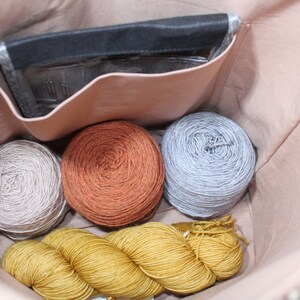May include: A light pink fabric bag interior containing three round yarn balls in beige, rust orange, and light gray, along with a long skein of mustard yellow yarn. A black rectangular pouch with a clear window holding needles is in the pocket.