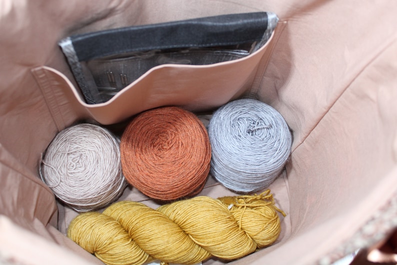 May include: A light pink fabric bag interior containing three round yarn balls in beige, rust orange, and light gray, along with a long skein of mustard yellow yarn. A black rectangular pouch with a clear window holding needles is in the pocket.