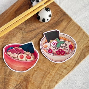Könnte beinhalten: Zwei illustrierte Aufkleber von Schüsseln mit Ramen. Eine Schüssel ist rot mit einem Seetang-Garnierung und die andere ist weiß mit einer grünen Garnierung.