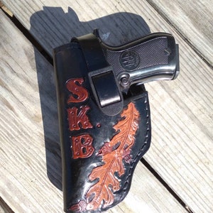 K&ouml;nnte beinhalten: Schwarze Lederholster mit einer Beretta-Pistole. Die Holster hat die Initialen "SKB" in roter Schrift und ein Eichenblatt-Design in Braun und Orange. Die Pistole ist silber und schwarz.