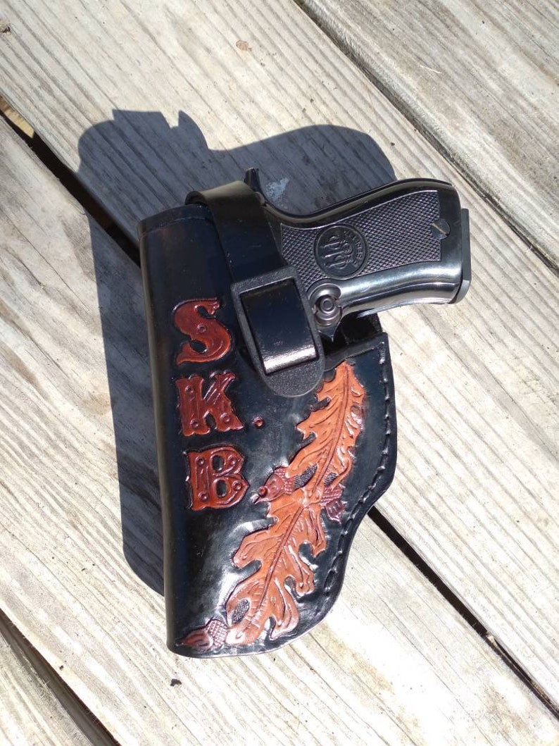 K&ouml;nnte beinhalten: Schwarze Lederholster mit einer Beretta-Pistole. Die Holster hat die Initialen "SKB" in roter Schrift und ein Eichenblatt-Design in Braun und Orange. Die Pistole ist silber und schwarz.